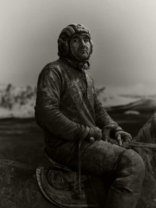 A man in muddy, rugged clothing and a helmet sits on a horse, gazing into the distance with a serious expression. Snow-covered mountains are blurred in the background.