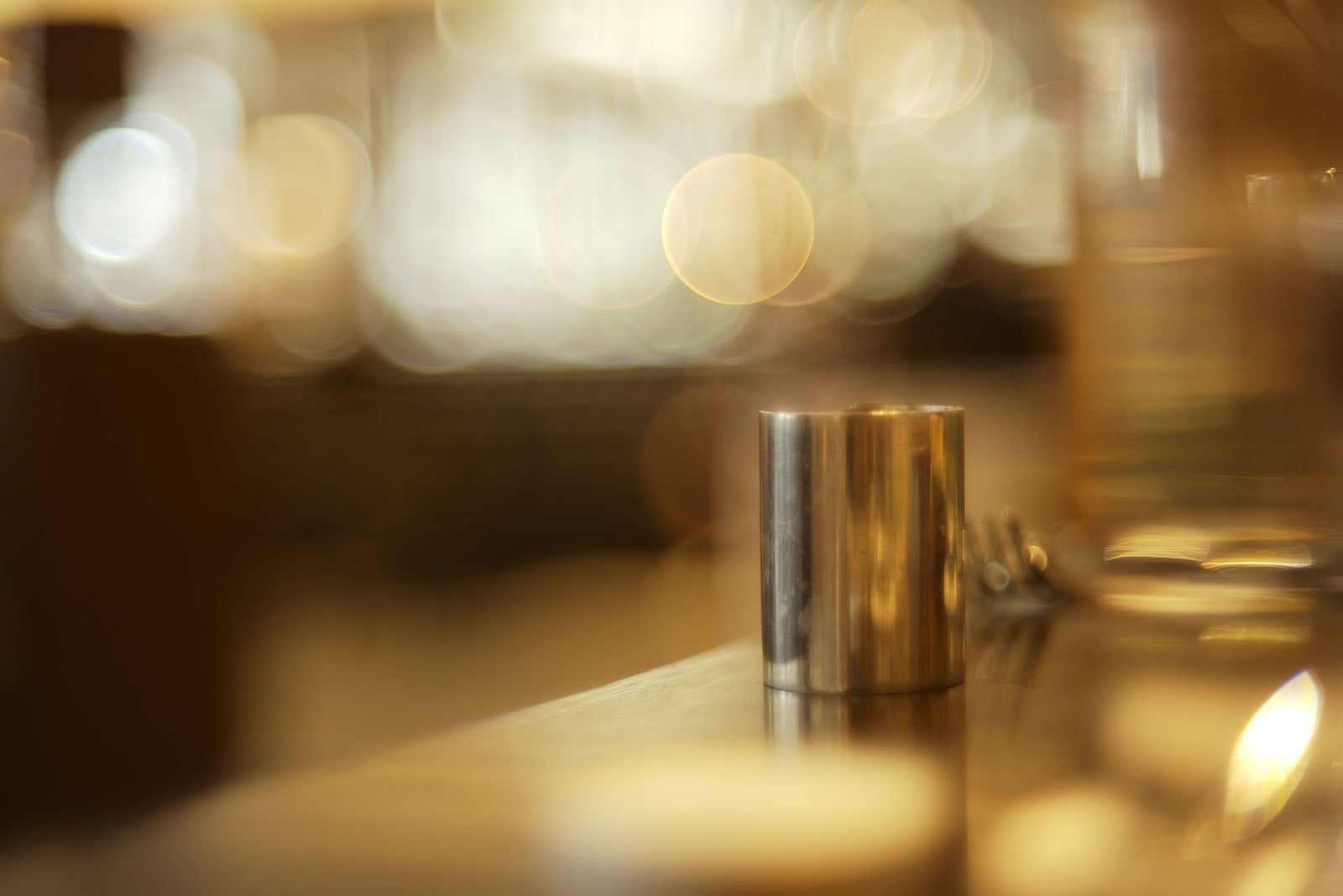 A small metal cylinder sits on a wooden surface with a blurred, warmly lit background featuring soft, circular light bokeh.