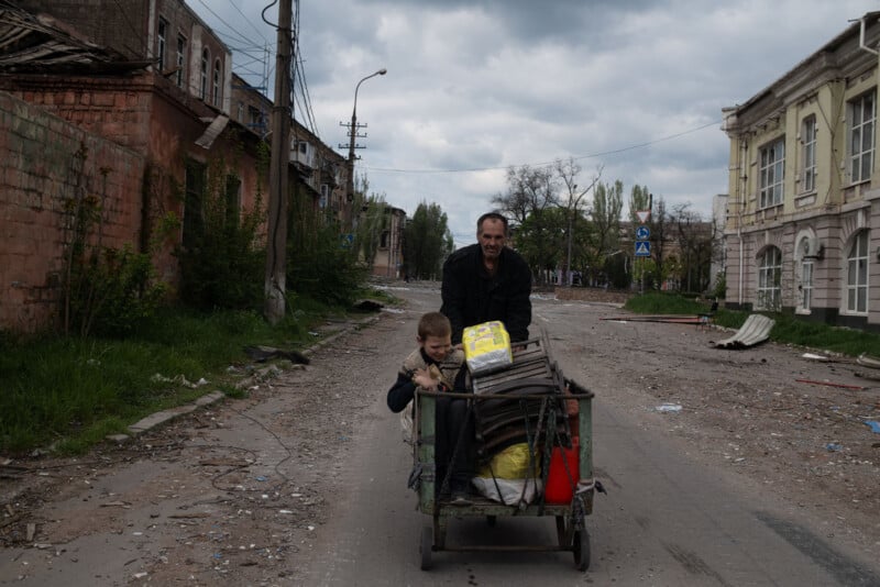 A man pushes a cart with a child and belongings down a damaged, debris-strewn street lined with dilapidated buildings under a cloudy sky.