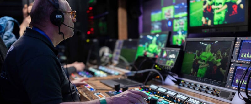 A person wearing headphones operates a control panel in a broadcast studio, with multiple monitors displaying live video feeds of a sports event, surrounded by various buttons and screens.