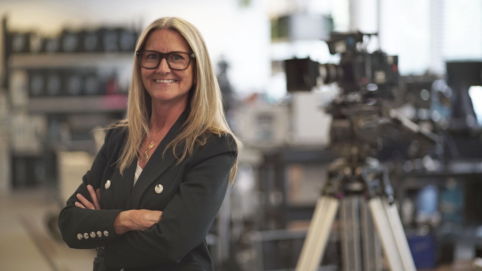 Smiling woman with long blonde hair and glasses stands with arms crossed in a professional setting, with a video camera on a tripod and shelves visible in the blurred background.