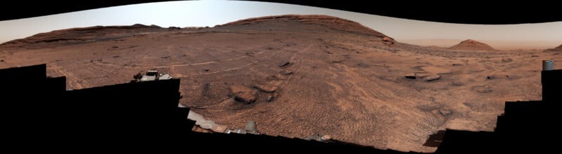 A panoramic view of a rocky, reddish Martian landscape with a large mountain in the distance and parts of a rover visible in the foreground.