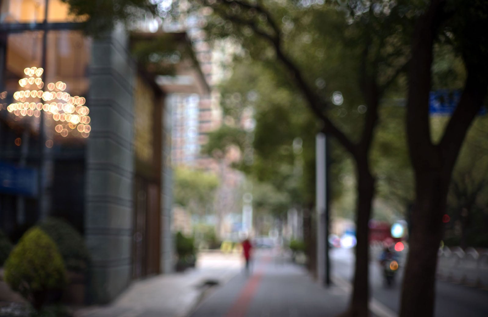 A city street scene taken out of focus, with blurred trees, buildings, a sidewalk, streetlights, and a chandelier in a window.