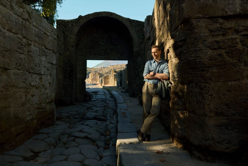 A person stands with arms crossed, leaning against an ancient stone wall on a sunlit cobblestone path, with ruins and an arched doorway visible in the background.