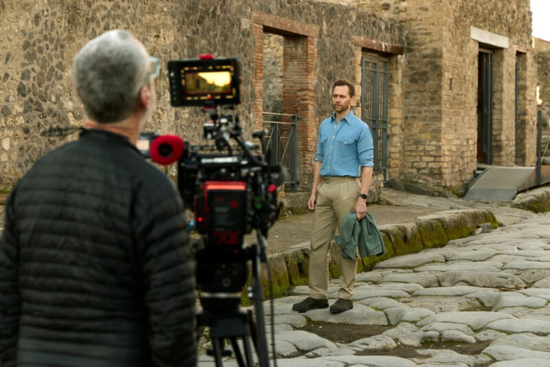 A cameraman films a man standing on ancient stone pavement in front of old brick buildings. The man holds a jacket and looks toward the camera, while the cameraman adjusts filming equipment.