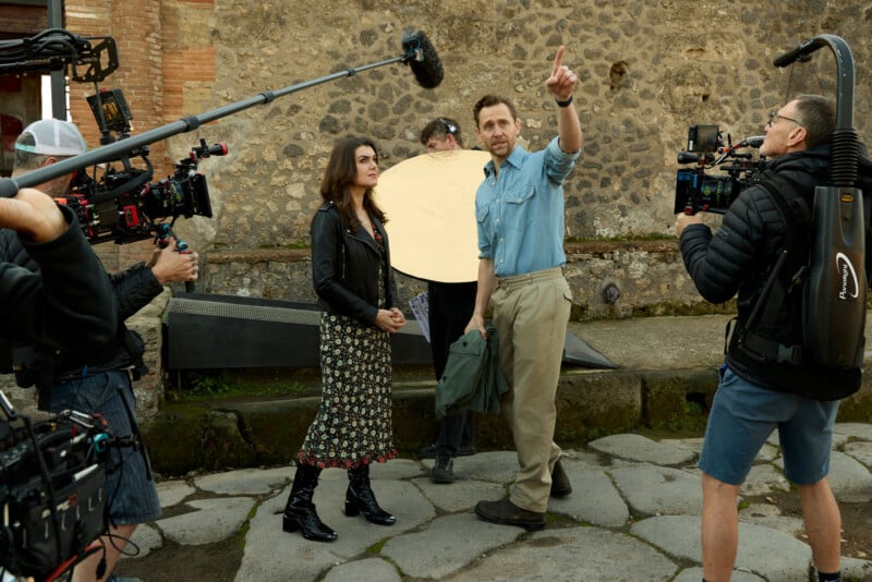 A film crew captures a man pointing while talking to a woman in a floral dress. Crew members hold cameras, a boom mic, and a reflector board. They stand on stone pavement against a textured stone wall.