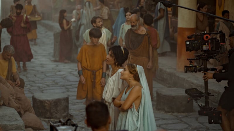Actors in ancient Roman-style costumes stand on a stone street film set, with camera equipment visible to the right and crew members capturing the scene.