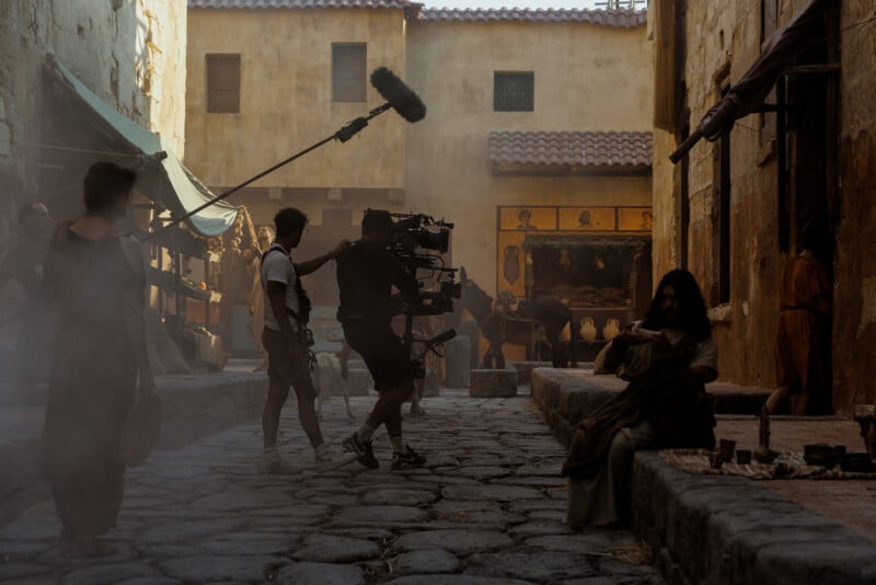 A film crew with a camera and boom mic records a scene on a cobblestone street set that resembles an ancient city, with actors in period costumes and dusty, golden lighting.