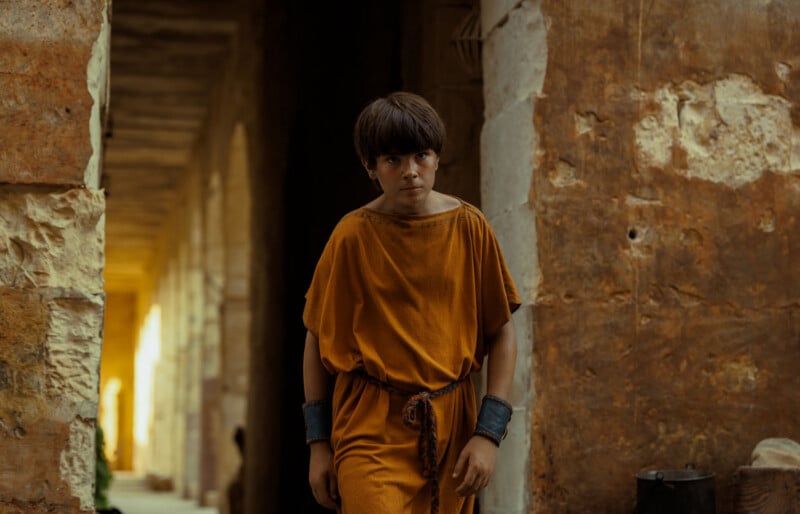 A young person in a loose orange tunic stands in a rustic stone corridor, looking forward with a serious expression. The warm-toned walls and arches suggest an ancient or historical setting.