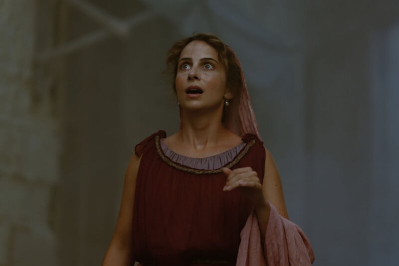A woman in a maroon dress and pink shawl looks upward with a surprised expression, standing against a blurred, grayish background.
