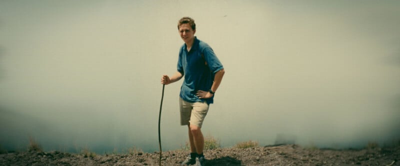 A person in a blue shirt and khaki shorts stands on rocky terrain, holding a walking stick, with fog or mist in the background.