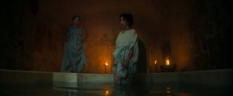 Two women in simple, loose dresses stand in a dim, ancient stone bathhouse lit by torches. One woman stands in the water, while the other stands on the edge, watching quietly.