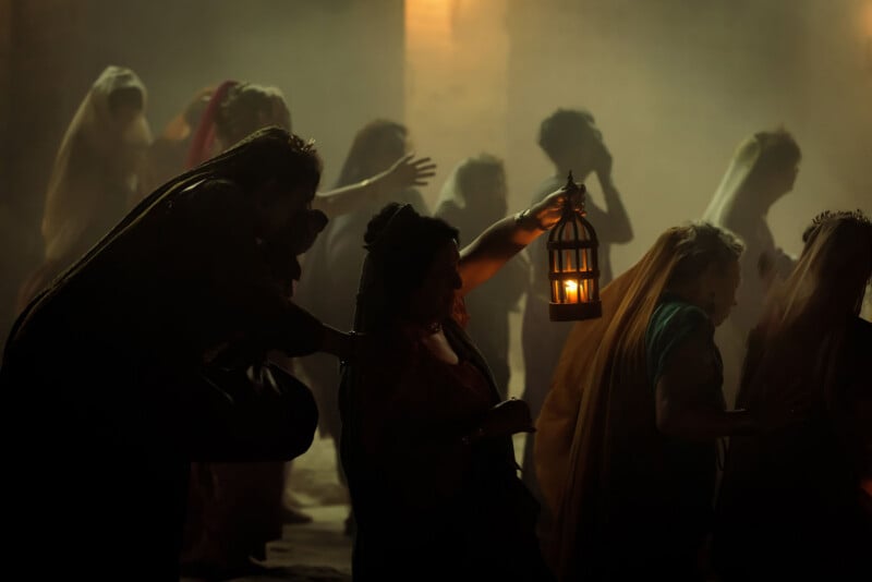 A group of people in robes and veils walk through a dim, misty setting. One person in the foreground holds up a lantern, illuminating their path. The scene appears historical or theatrical.