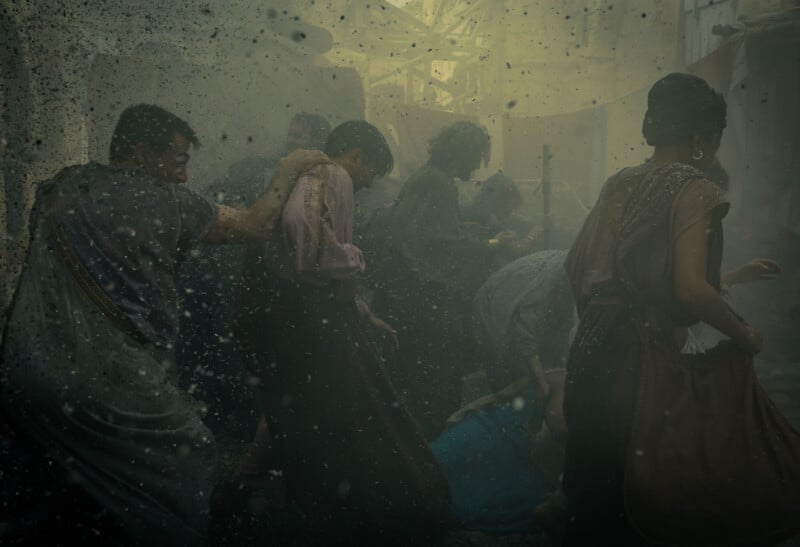 A group of people in historical clothing struggle through a chaotic, dusty scene, with debris and particles filling the air. Some are helping each other while others crouch or fall amid the turmoil.