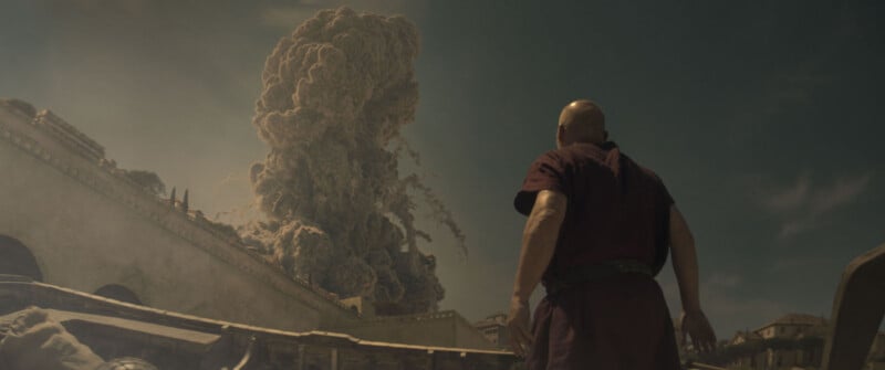 A person in ancient clothing looks toward a massive volcanic eruption, with a large ash cloud rising over buildings under a dark sky.