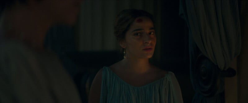A woman in a light blue dress with a red mark on her forehead stands indoors, looking toward the camera with a serious expression. Curtains and dim lighting surround her.