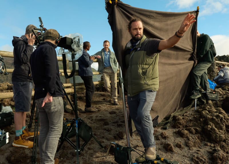A film crew works outdoors with cameras and equipment as an actor stands in front of a brown backdrop, while a crew member gestures with his arm extended.