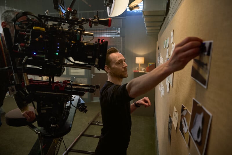 A man arranges photos on a bulletin board while a camera crew films him on a set, capturing the scene in a dimly lit room with props and lighting equipment visible.