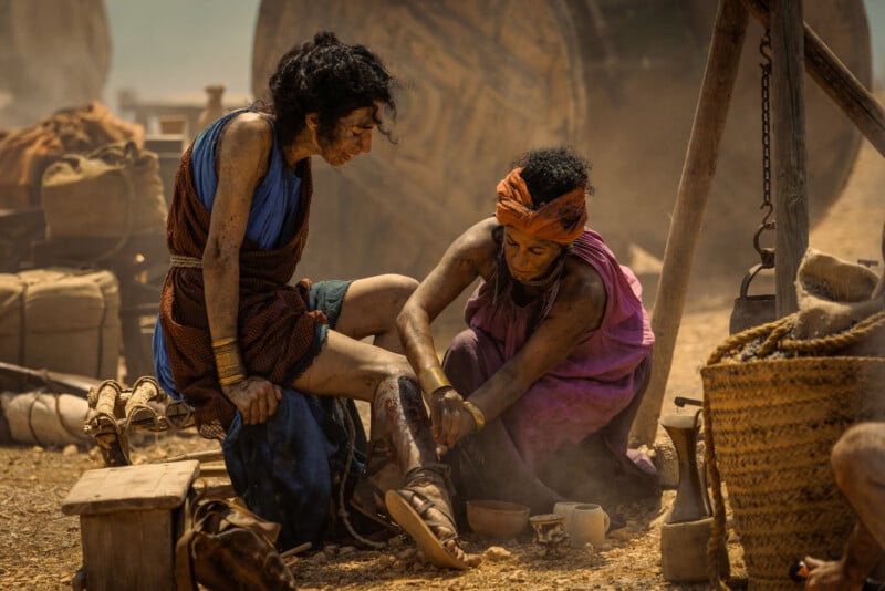 Two women in ancient-style clothing sit on the ground in a dusty, outdoor setting; one tends to the other's injured leg, wrapping it with cloth, surrounded by baskets, pottery, and large wooden wheels.