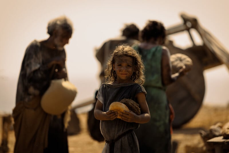 A young child with messy hair and dirt-stained face holds bread, standing in front of blurred figures and a wooden cart in a dusty, sepia-toned setting, suggesting a historical or ancient scene.
