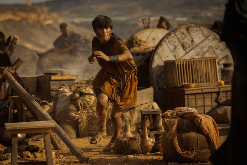 A young person in brown ancient-style clothing runs through a dusty, cluttered outdoor market with pottery, crates, and baskets scattered around in a sepia-toned scene.