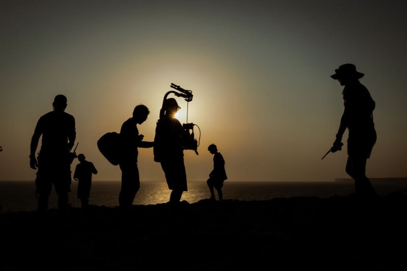 Silhouetted figures stand and walk along a coastline at sunset, with one person holding camera equipment. The sun sets over the water in the background.