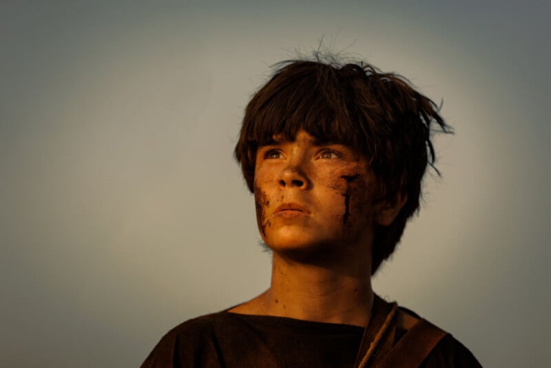 A young boy with short dark hair and a dirty face looks up with a determined expression, standing against a clear sky and wearing a dark shirt and strap.
