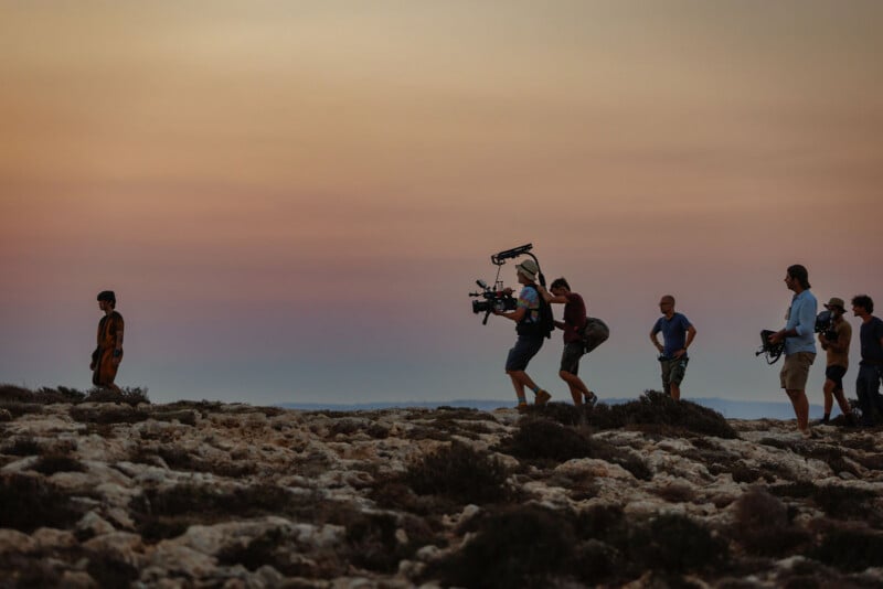 A film crew with cameras follows an actor walking across a rocky landscape at sunset, silhouetted against a colorful sky.