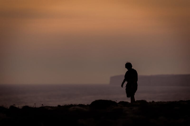 A silhouette of a person walking along a coastline at sunset, with calm water and distant cliffs under an orange and purple sky.