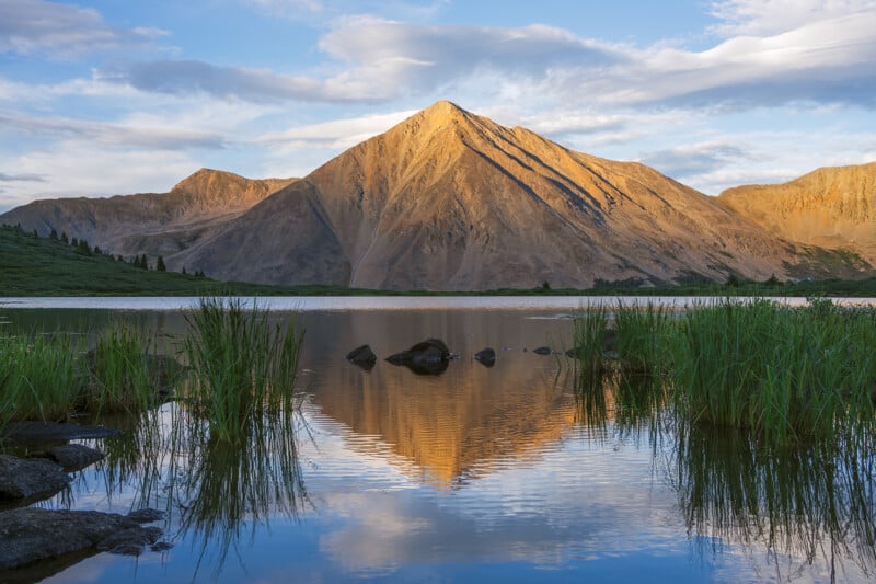 A sunlit mountain reflects in a calm lake surrounded by green reeds, under a blue sky with scattered clouds.