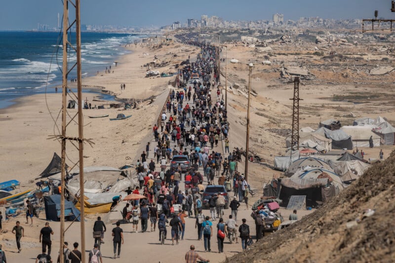 A large crowd of people walks along a coastal road between the sea and damaged tents, with buildings and ruins visible in the background under a clear sky.