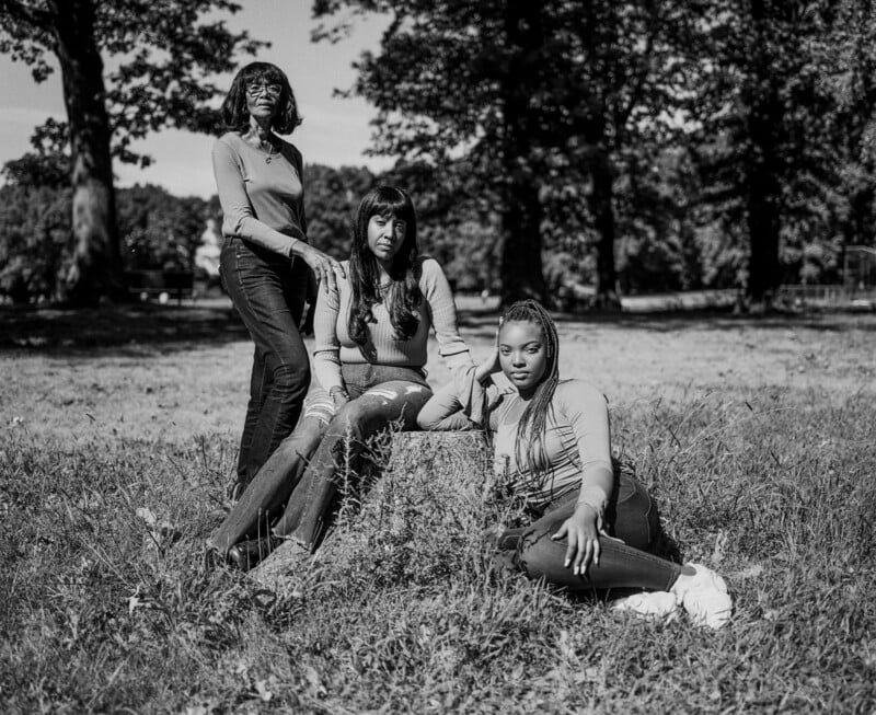 Three women pose together in a grassy, sunlit park with tall trees in the background. Two women sit on the grass while one stands behind them, all looking toward the camera. The photo is in black and white.