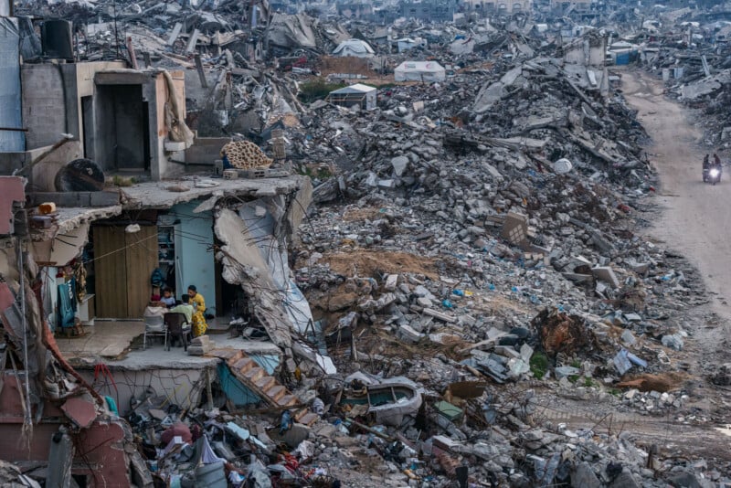 A family sits at a table in a partially destroyed building, surrounded by rubble and debris, with a dirt road and more destruction in the background.