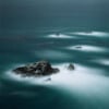 Rocky outcrops rise from a turquoise sea, partially shrouded by mist. The water appears smooth and hazy, creating a dreamlike, tranquil atmosphere under a soft, overcast sky.