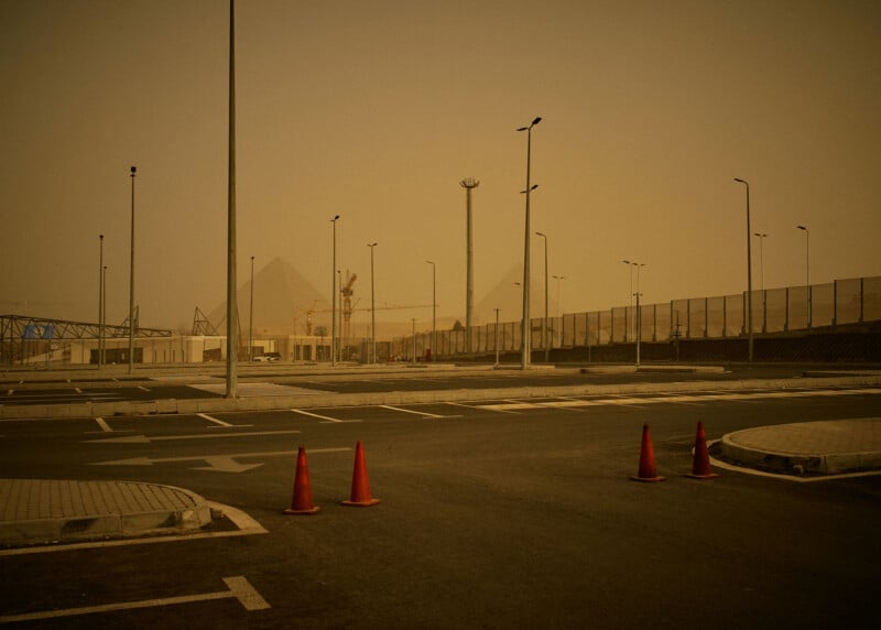 An empty, hazy parking lot with red cones blocking a lane; tall light poles line the area, and pyramids are faintly visible in the background through the dust.
