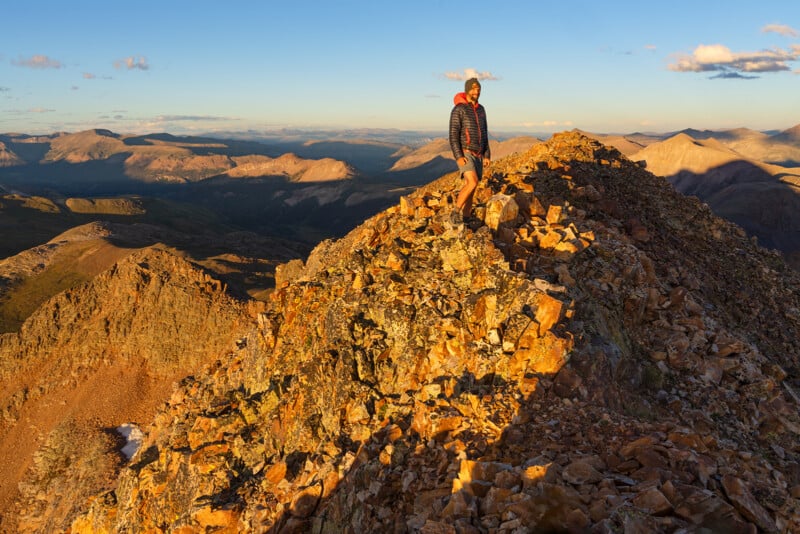 A person in outdoor clothing stands on a rocky mountain ridge at sunset, surrounded by dramatic peaks and a colorful sky, with warm sunlight illuminating the landscape.