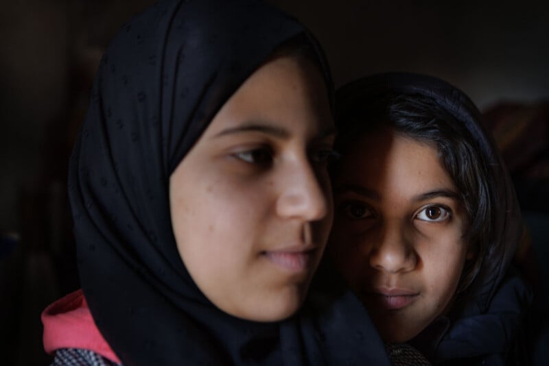 Two girls, one in a dark hijab and the other with loose dark hair, stand close together. The light softly illuminates their faces, with one girl's eyes looking directly at the camera, while the other looks away.
