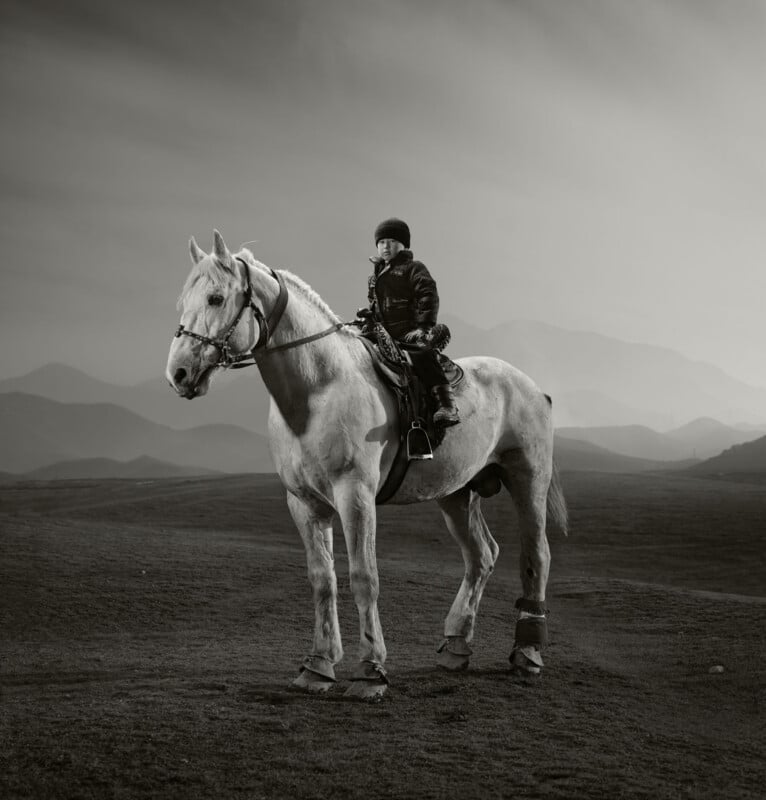 A person wearing a helmet and jacket sits atop a white horse on an open plain, with distant mountains and a cloudy sky in the background. The image is in black and white.