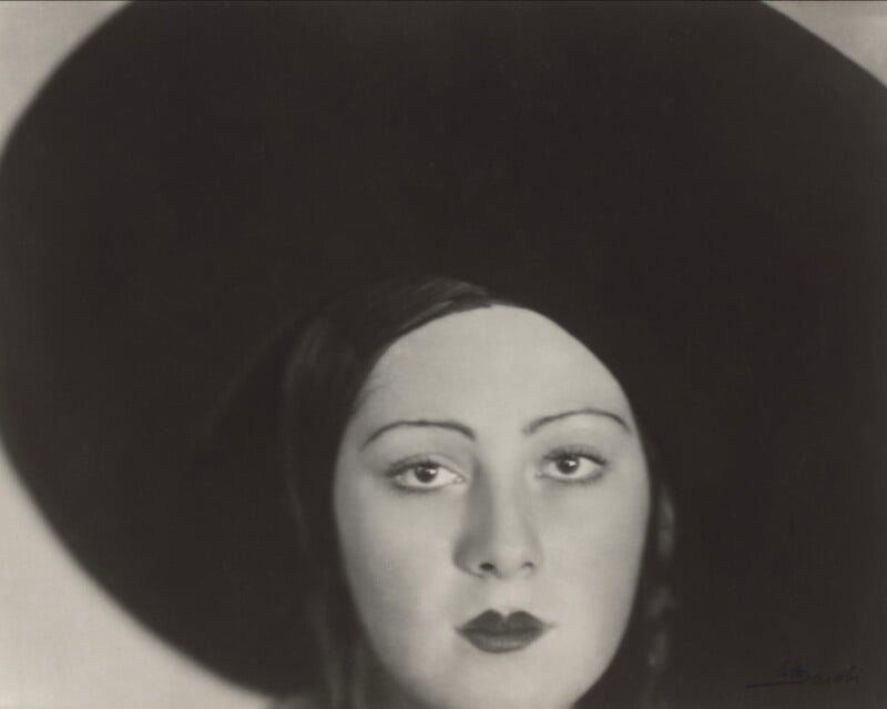 A close-up, black-and-white portrait of a woman wearing a large, dark, wide-brimmed hat that frames her face.