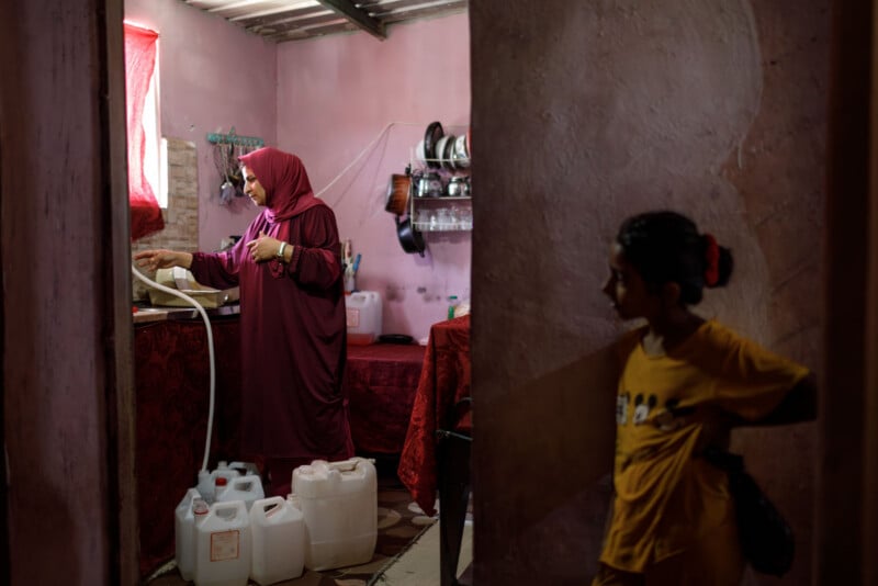 A woman in a maroon dress fills a jug with water in a small pink kitchen, while a child in a yellow shirt stands in a dimly lit doorway, looking inside. Several white water containers are on the floor.