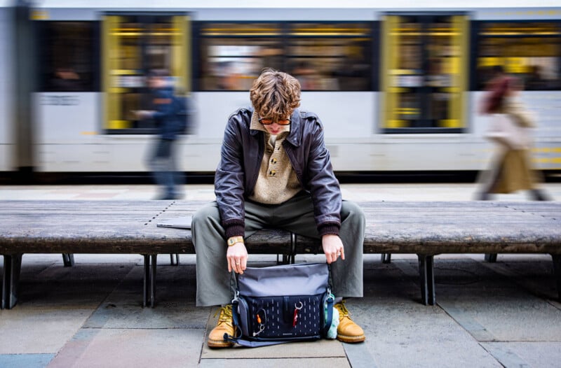 A person sits on a bench with their head down, looking into an open bag on their lap. People and a tram move quickly in the blurred background, suggesting a busy urban setting.