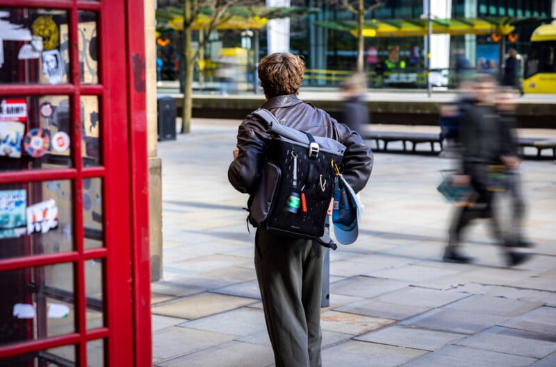 A person with short hair and a leather jacket walks on a city sidewalk, carrying a backpack with a water bottle. A red phone booth with stickers is on the left, and blurred pedestrians and a yellow tram are in the background.