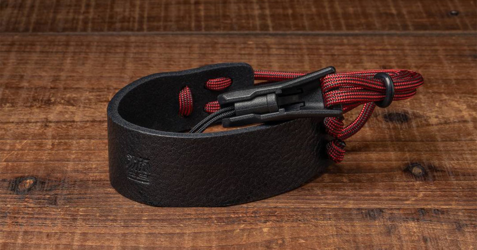 A black leather wristband with a black plastic buckle and red paracord detail is displayed on a wooden surface.