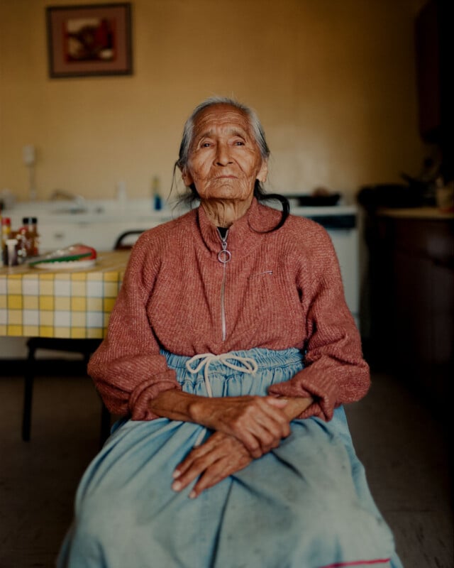 An elderly woman with long gray hair sits indoors, wearing a reddish sweater and blue skirt. She has her hands folded in her lap and looks calmly ahead. A table with a checkered tablecloth is visible behind her.