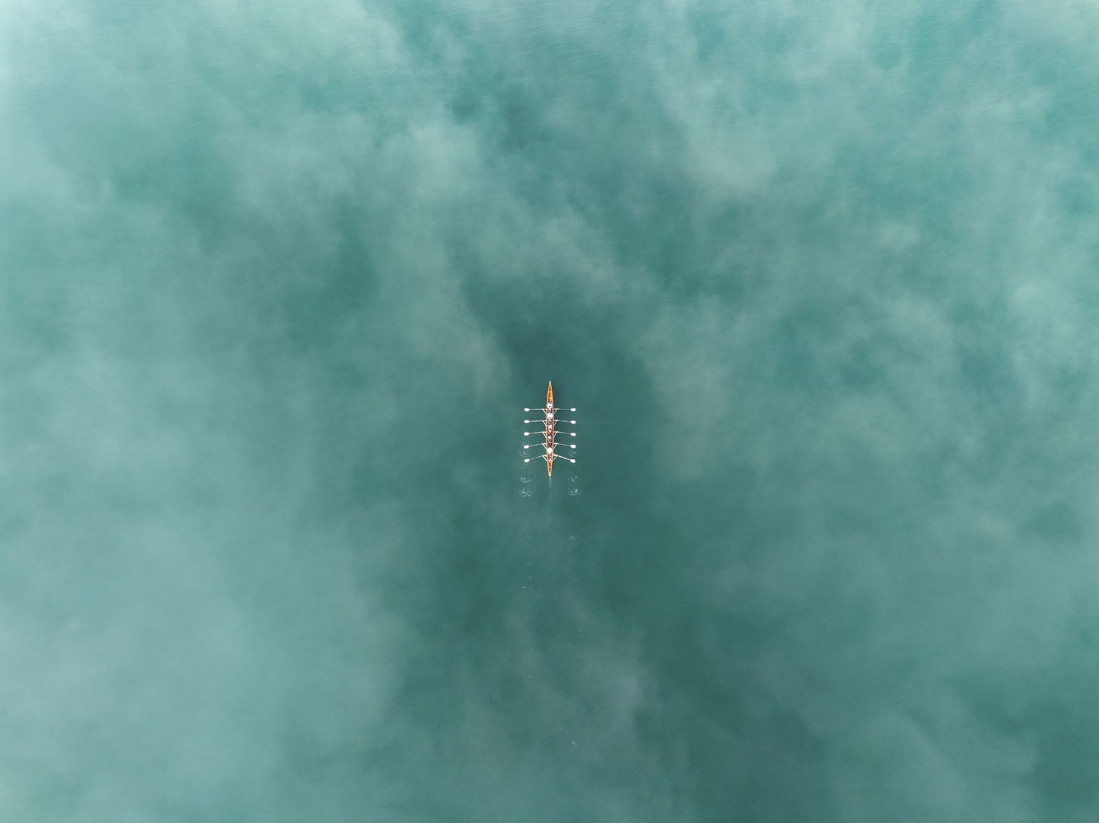 Aerial view of a rowing team in a narrow boat on calm, turquoise water, with oars creating small ripples, surrounded by mist or fog.