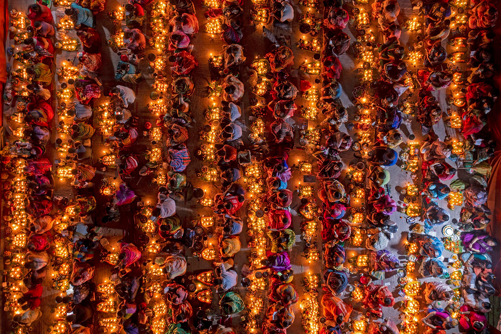 A vibrant overhead view of a large group of people sitting in rows, each surrounded by glowing candles, creating a colorful and festive atmosphere during a traditional celebration or festival.