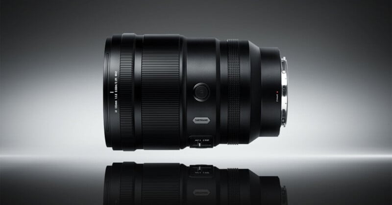 A professional camera lens is displayed horizontally against a gradient gray background, with its reflections visible beneath it. The lens features various controls and textured grip rings.