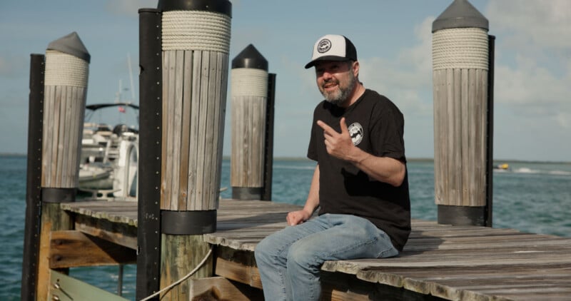 A man in a black t-shirt and cap sits on a wooden dock by the water, gesturing with his hand. Several boats are moored in the background under a partly cloudy sky.