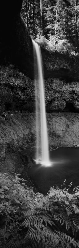 A tall, thin waterfall plunges from a rocky cliff into a dark pool, surrounded by a lush forest and a walking path.