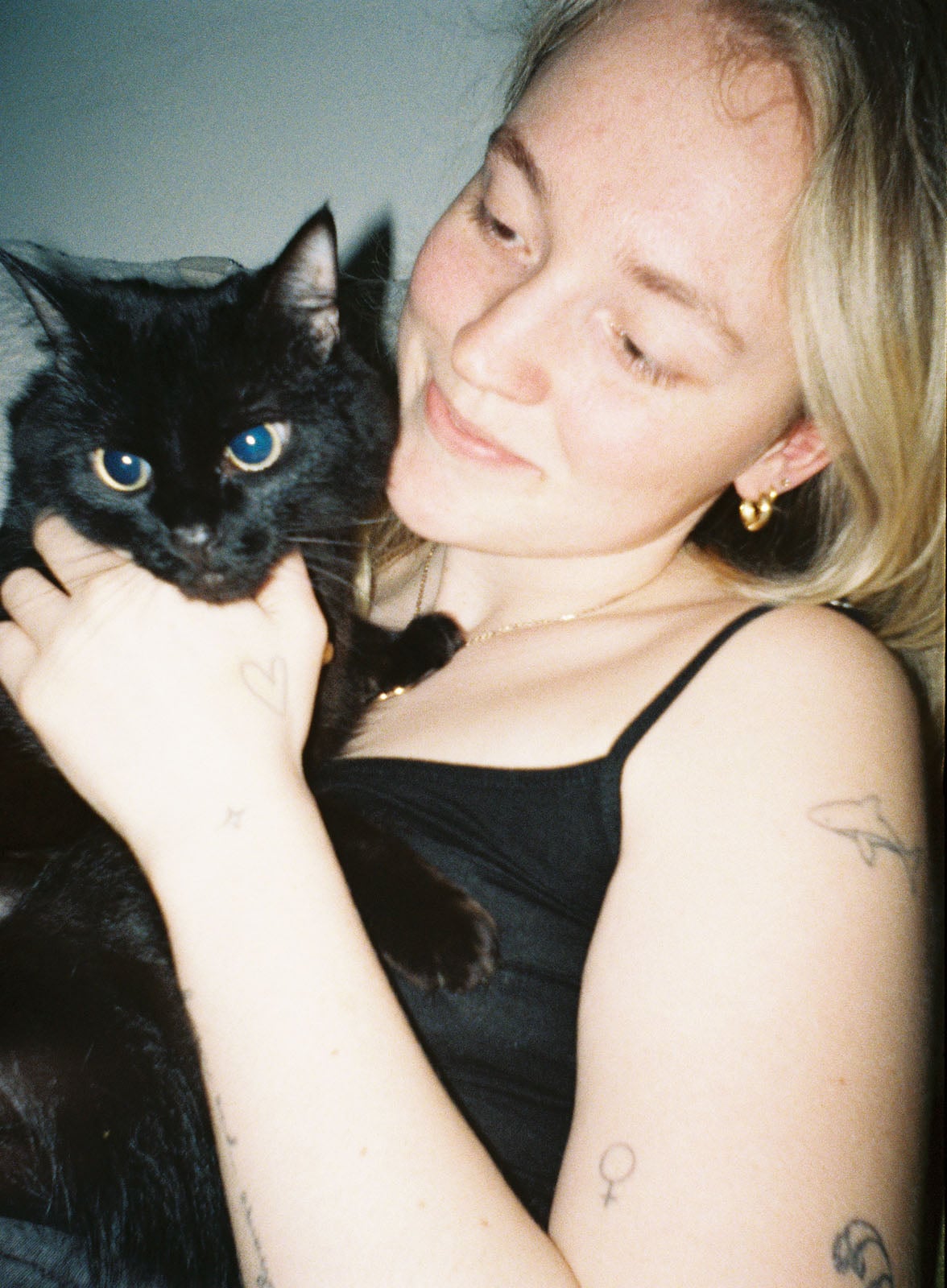 A smiling person with light hair holds a fluffy black cat. The person has several tattoos on their arms, including a heart, star, planet, and Venus symbol. Both are looking slightly away from the camera.